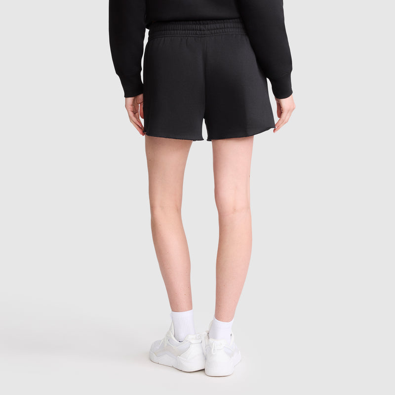A person is shown from behind, wearing a black sweatshirt and comfortable athletic shorts—specifically Champion’s 3" Powerblend Shorts, C Logo—paired with white socks and white sneakers, set against a plain light background.