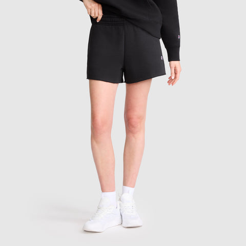 A person is wearing the Champion 3" Powerblend Shorts, C Logo, paired with white socks and white sneakers. Standing against a plain light background, only the lower half of their body is visible. One hand rests on the waistband to highlight these comfortable athletic shorts from Champion.