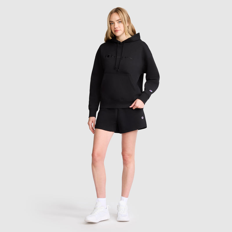 A woman stands against a plain light gray background, wearing a black hoodie, white sneakers, and Champion 3" Powerblend Shorts with the C Logo. She has blonde hair and a relaxed posture, with one hand in her pocket.