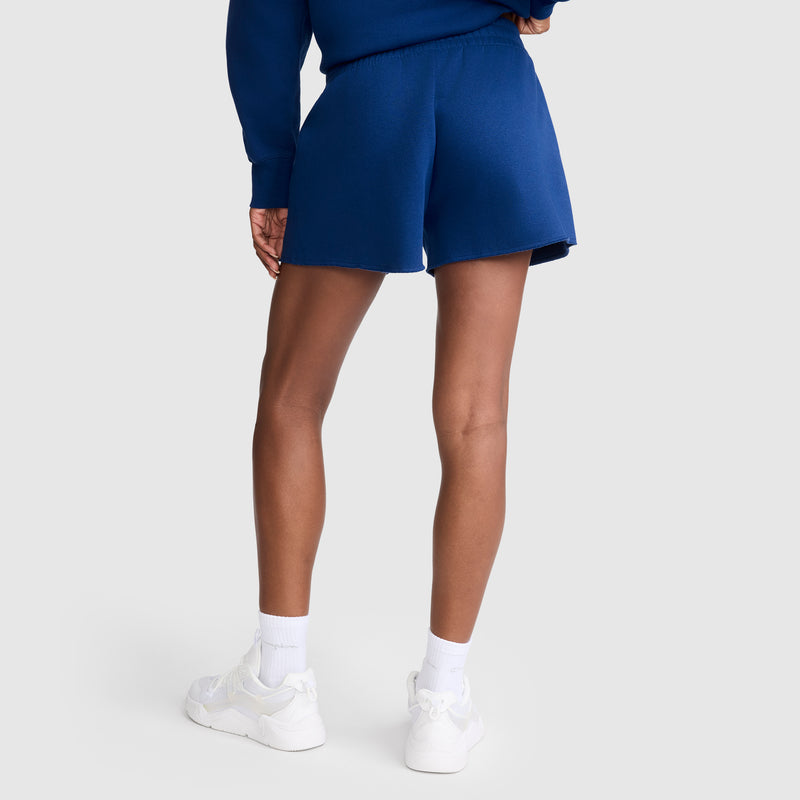 A person stands with their back to the camera against a plain light background, wearing a blue sweatshirt paired with Champion 3" Powerblend Shorts featuring the signature C Logo, along with white socks and white sneakers.