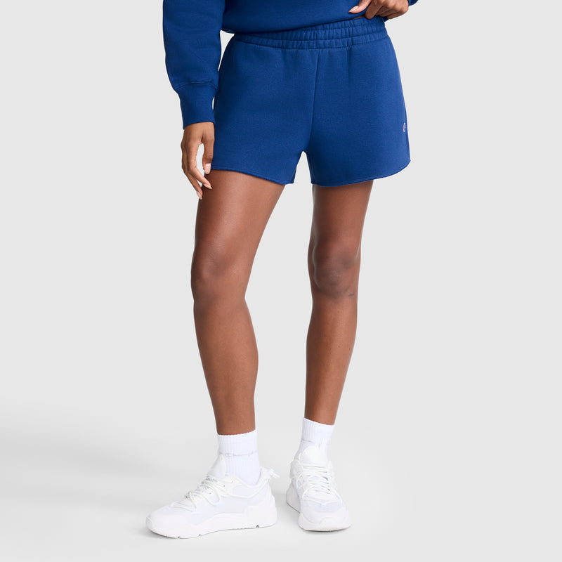 A person is shown from the waist down wearing Champion's 3" Powerblend Shorts with the iconic C Logo in blue, paired with a matching blue sweatshirt, white crew socks, and white sneakers, standing against a plain light gray background. The comfortable athletic Champion shorts make this look perfect for casual or sporty outfits.