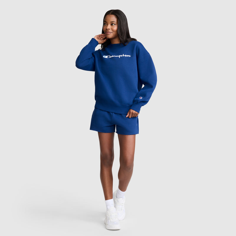 A woman stands smiling with one hand touching her face against a plain light grey background, wearing a blue Champion sweatshirt, Champion 3" Powerblend Shorts, C Logo, and white sneakers. Her comfortable athletic shorts from Champion perfectly complement her casual look.