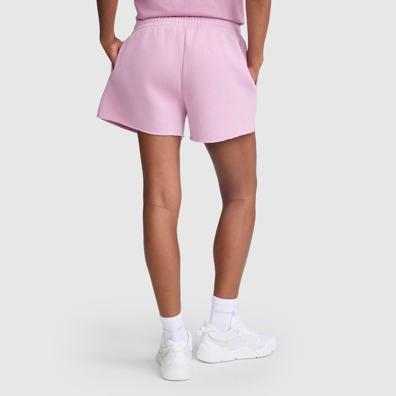 A person is shown from the back on a plain light gray background, wearing the Champion 3" Powerblend Shorts, C Logo in light pink along with a matching shirt, white crew socks, and white sneakers, standing with hands in the shorts' pockets.