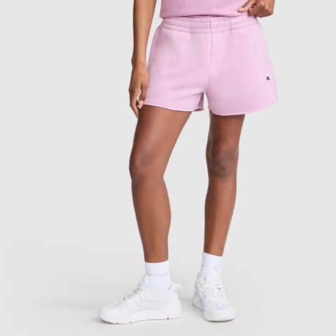 A person wears Champion 3" Powerblend Shorts, C Logo in light pink, featuring a small blue and red Champion logo on the left leg. These comfortable athletic shorts are paired with white crew socks and white sneakers, set against a light gray background.