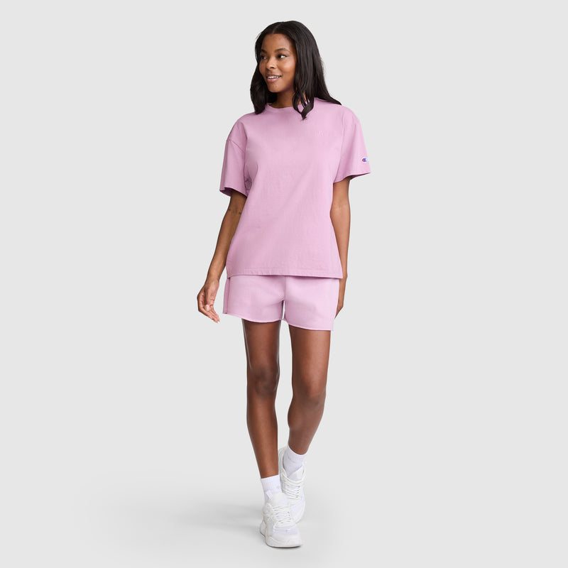 A woman wearing Champion’s 3" Powerblend Shorts, C Logo in light pink and a matching t-shirt smiles while walking. She has shoulder-length hair and completes her look with white socks and sneakers, standing against a plain light gray background.