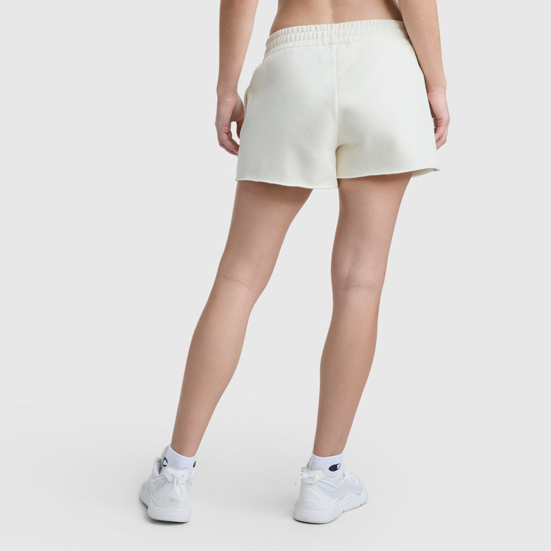A person stands facing away, wearing Champion 3" Powerblend Shorts with C Logo, a cropped top, white athletic socks, and white sneakers against a plain light background.