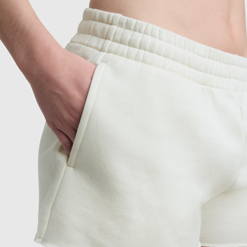 Close-up of a person wearing Champion 3" Powerblend Shorts, C Logo—off-white, high-waisted women’s shorts with an elastic waistband and a hand in the pocket. Background is plain and light-colored.