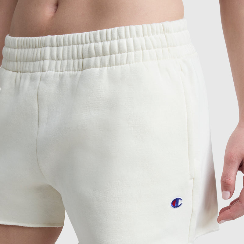 A close-up of a person wearing white Champion 3" Powerblend Shorts, C Logo, featuring an elastic waistband and embroidered Champion C logo near the left hem. The person stands against a plain light background.