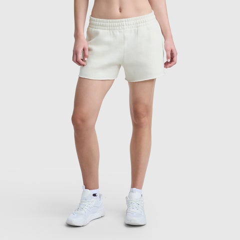 Someone models Champion's 3" Powerblend Shorts with the C Logo, paired with a bare midriff and white athletic sneakers, standing against a plain white background. The face is not shown.