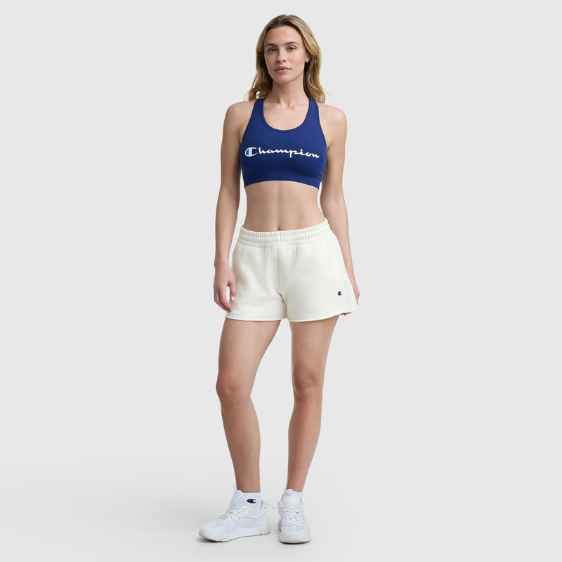 A woman faces forward, wearing a blue Champion sports bra and cream-colored 3" Powerblend Shorts, C Logo by Champion, paired with white socks and sneakers, against a plain light gray background.