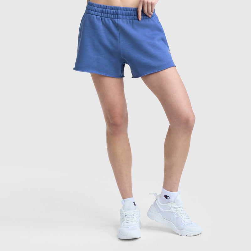 A person wears blue Champion 3" Powerblend Shorts, C Logo, with white crew socks and white sneakers, standing against a plain light background. Only their lower body is visible.
