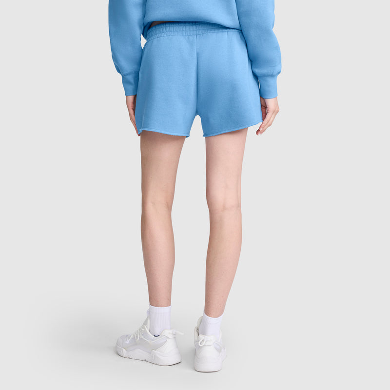 A person is standing facing away, wearing a blue sweatshirt and a pair of Champion 3" Powerblend Shorts with the C Logo, paired with white socks and white sneakers, set against a plain light background.