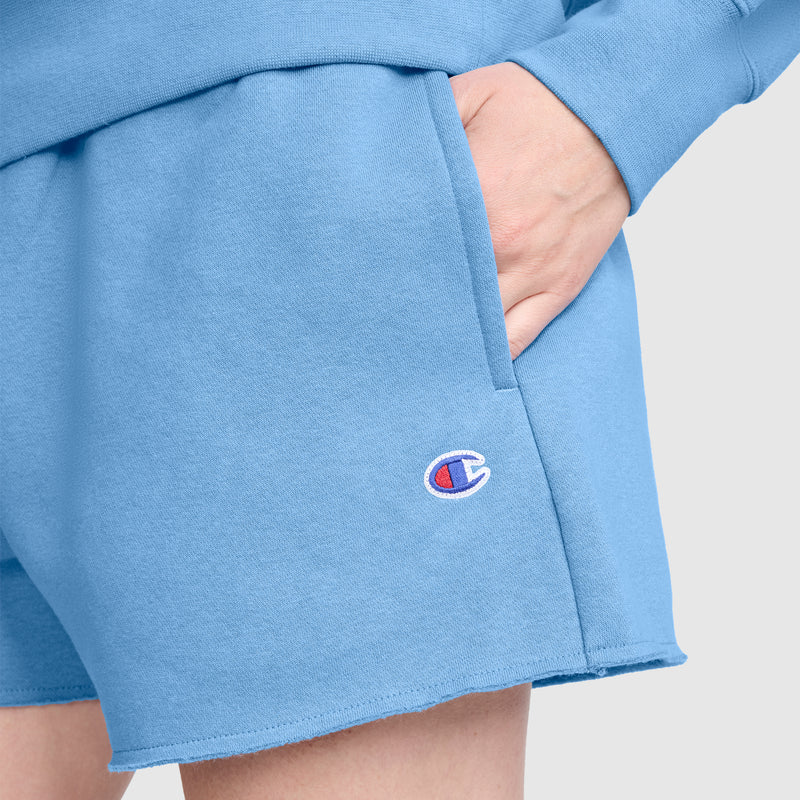A person wearing light blue Champion 3" Powerblend Shorts, C Logo with their hand in the pocket. These comfortable athletic shorts feature the Champion logo embroidered near the hem.
