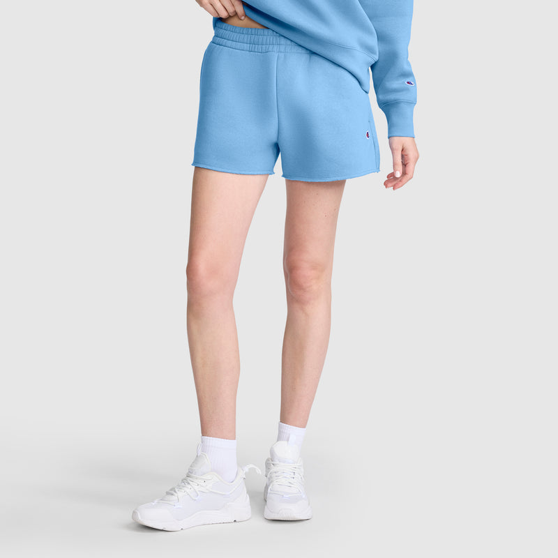 A person is wearing Champion’s 3" Powerblend Shorts, C Logo in light blue, paired with a matching sweatshirt, white socks, and white sneakers. They are standing against a plain light background with their left hand resting on their hip.