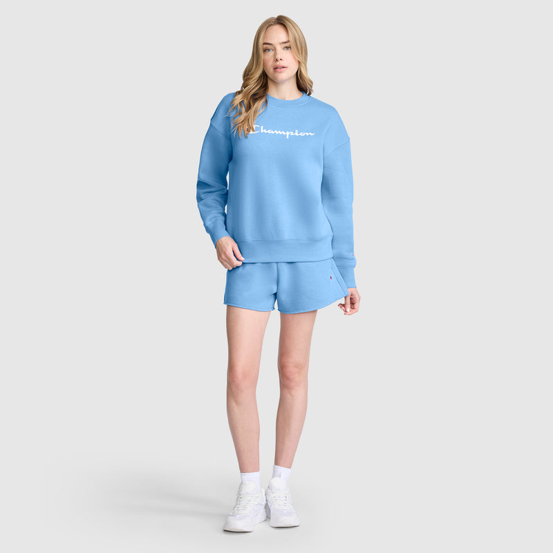 A person stands against a plain light gray background wearing a matching light blue sweatshirt and Champion 3" Powerblend Shorts featuring the signature C Logo, paired with white socks and white sneakers.