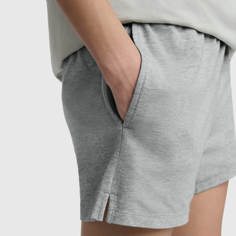 A person wearing light gray Champion Practice Shorts with a hand in the pocket. These comfy shorts feature a slight hem slit and are paired with a light-colored top.