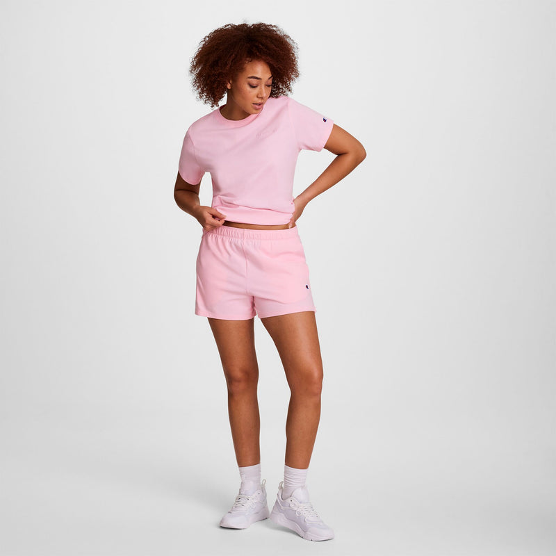 A woman with curly hair stands against a white background, wearing a light pink T-shirt, Champion Practice Shorts, white sneakers, and socks. She looks down with one hand on her waist.