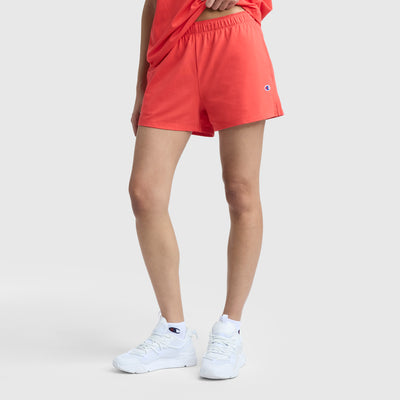 A person wears Champion’s coral Practice Short with a matching top and white sneakers with blue accents, posing against a light gray background. The sporty outfit features Champion logos throughout.