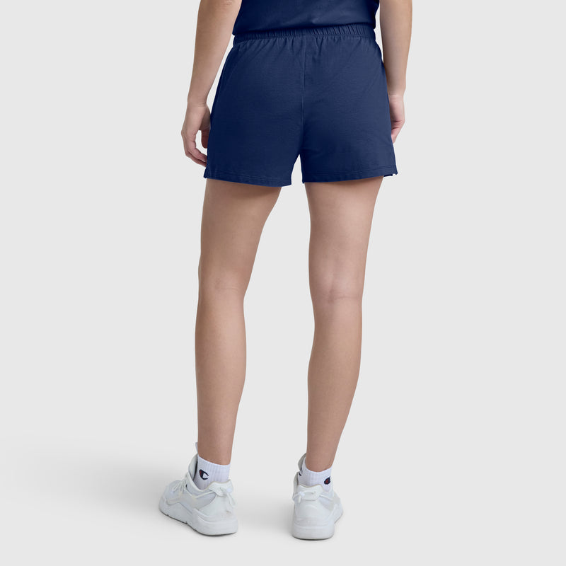 A person stands facing away, dressed in a navy blue T-shirt and Champion Practice Short, paired with white socks and white sneakers against a plain light background.