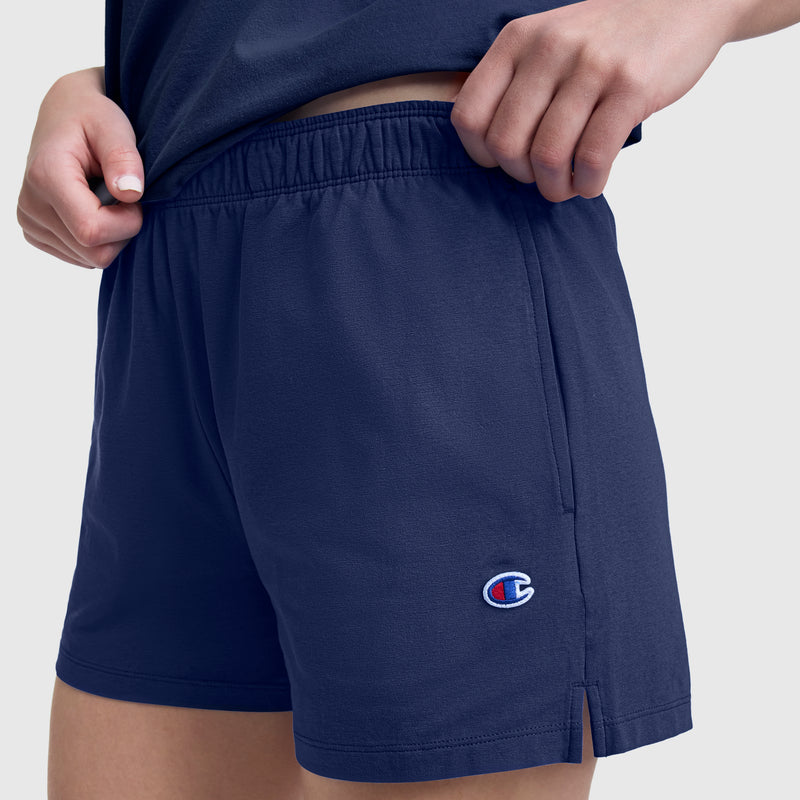 A person wearing Champion Practice Shorts in navy blue, featuring an elastic waistband and a small Champion logo near the hem, while holding up the hem of their matching navy shirt.