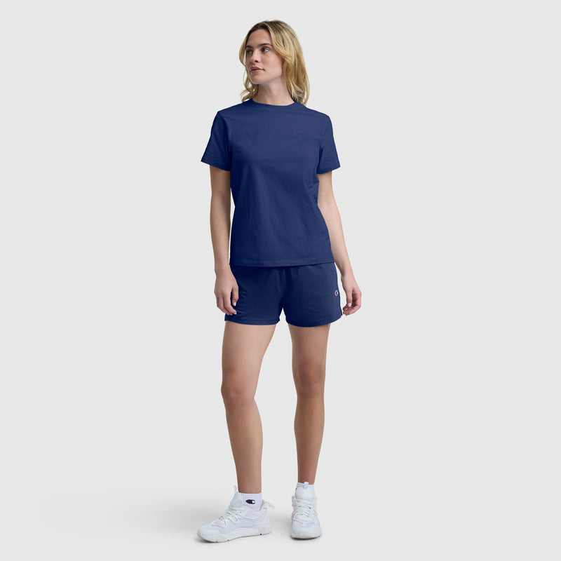 A person stands against a plain background wearing a navy blue T-shirt, Champion Practice Shorts in matching navy blue, and white sneakers. They have shoulder-length blonde hair and a neutral expression, with one arm relaxed and the other slightly bent.