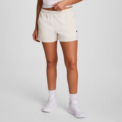 Wearing a white top, Champion Practice Short in cream, white socks, and sneakers, this person stands against a plain light gray background with hands at their sides and head out of frame—perfect comfy shorts for women.