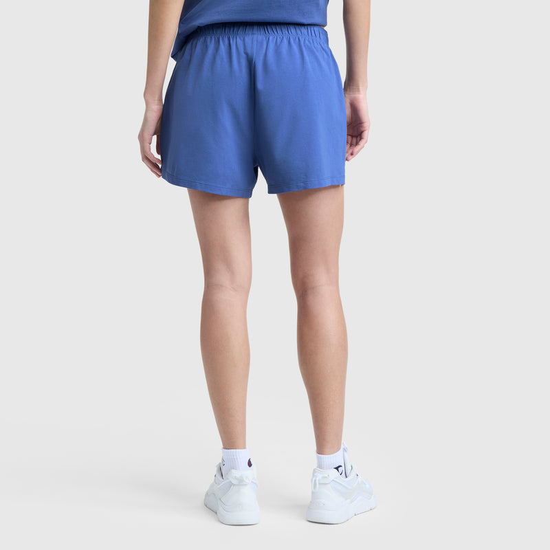 A person faces away, showing only their lower back and legs, wearing a blue shirt, Champion Practice Short with an elastic waistband, white crew socks, and white sneakers against a light gray background.