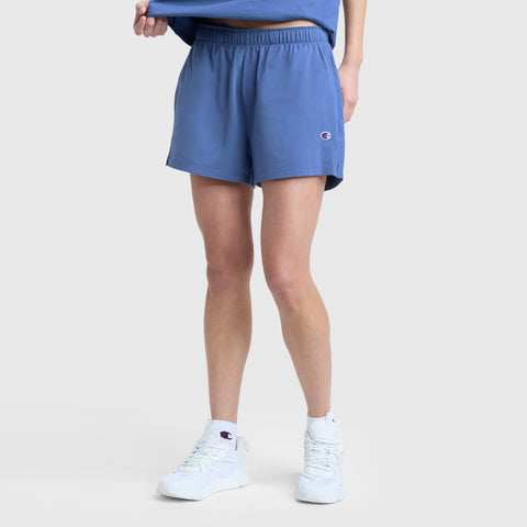 A person is shown from the waist down, wearing Champion Practice Shorts in light blue with an elastic waistband, paired with a matching blue shirt and white sneakers, standing against a plain light gray background.