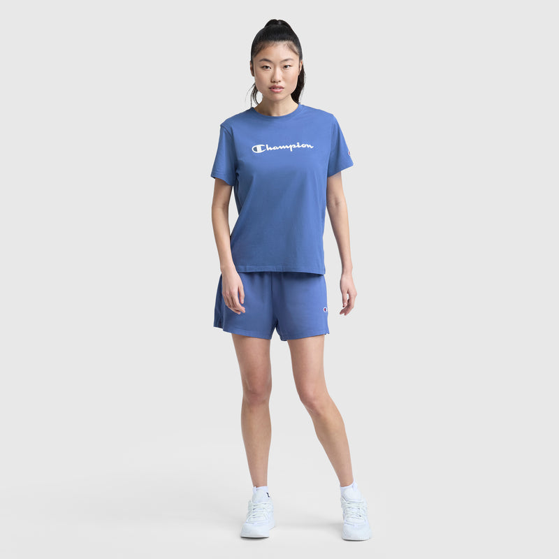 A woman with long dark hair in a high ponytail stands against a white background, wearing Champion's blue Practice Short with an elastic waistband and matching Champion t-shirt, looking directly at the camera.