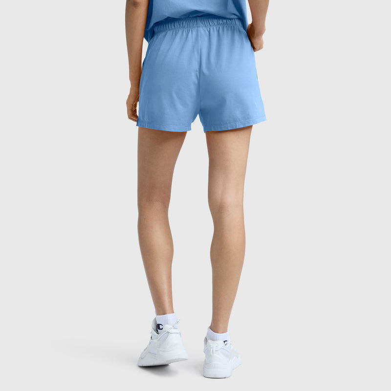 A person stands with their back to the camera, wearing a light blue T-shirt, Champion Practice Shorts in a matching light blue shade, and white sneakers against a plain background.