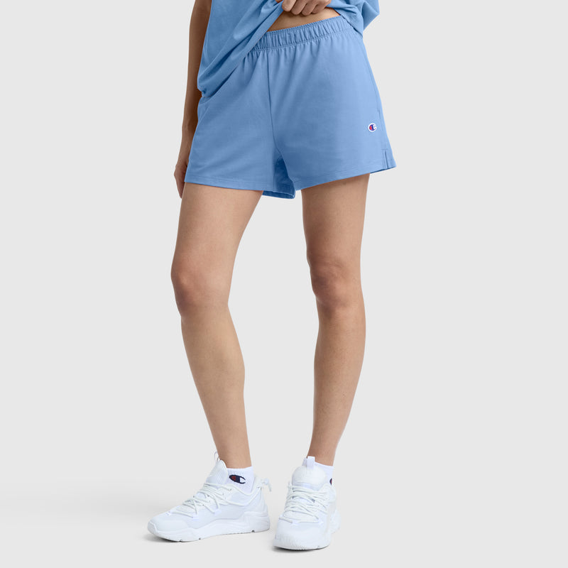 A person wearing Champion’s Practice Short in light blue, paired with a matching t-shirt and white athletic sneakers, stands against a plain light gray background.