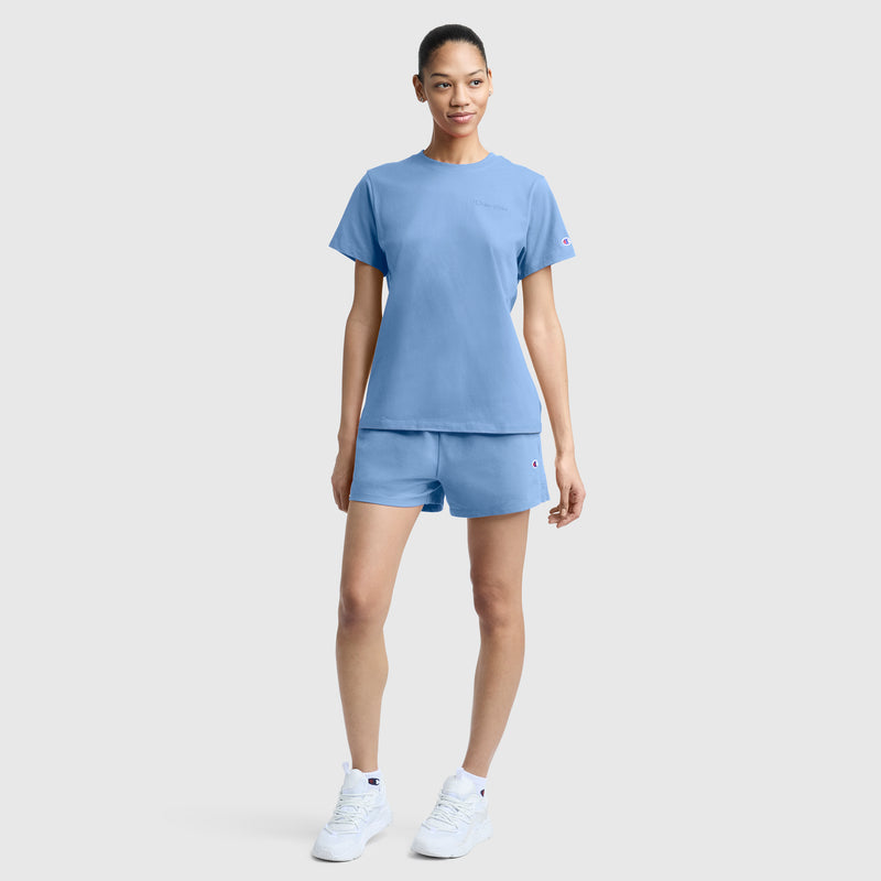 A woman stands against a plain background, wearing the Champion Practice Short in light blue paired with a matching T-shirt and white sneakers. Her hair is pulled back, and she smiles slightly with one hand by her side and the other relaxed.