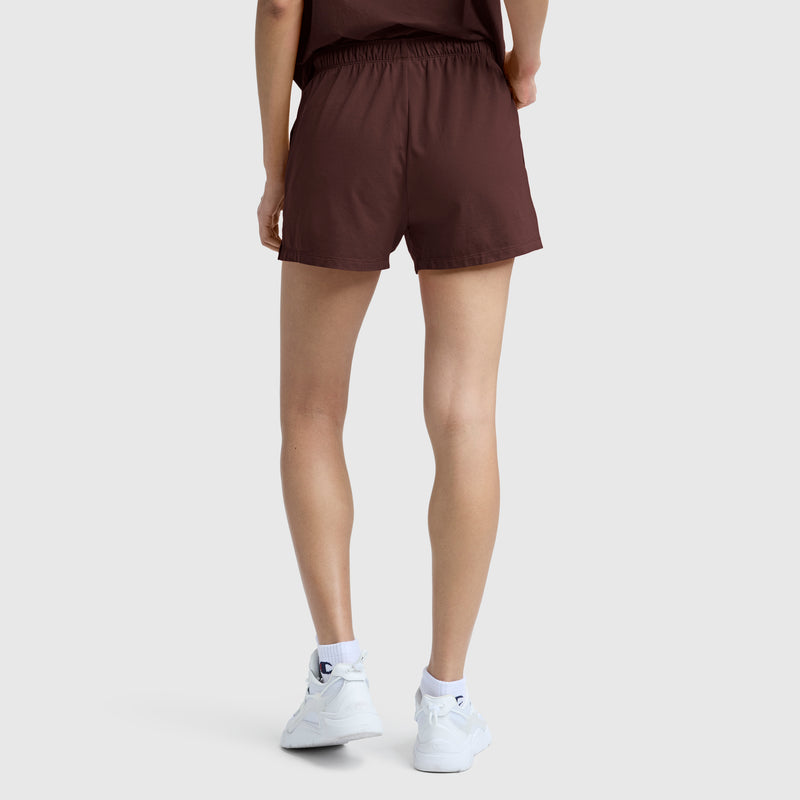 A person stands facing away, wearing a brown t-shirt and the Champion Practice Short—slim-fit women’s shorts made from a comfortable cotton blend—paired with white sneakers and socks, against a plain light background.