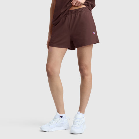 A person is shown from the waist down, standing against a plain light gray background, wearing maroon Champion Practice Shorts featuring a small logo, paired with a matching maroon t-shirt and white sneakers.