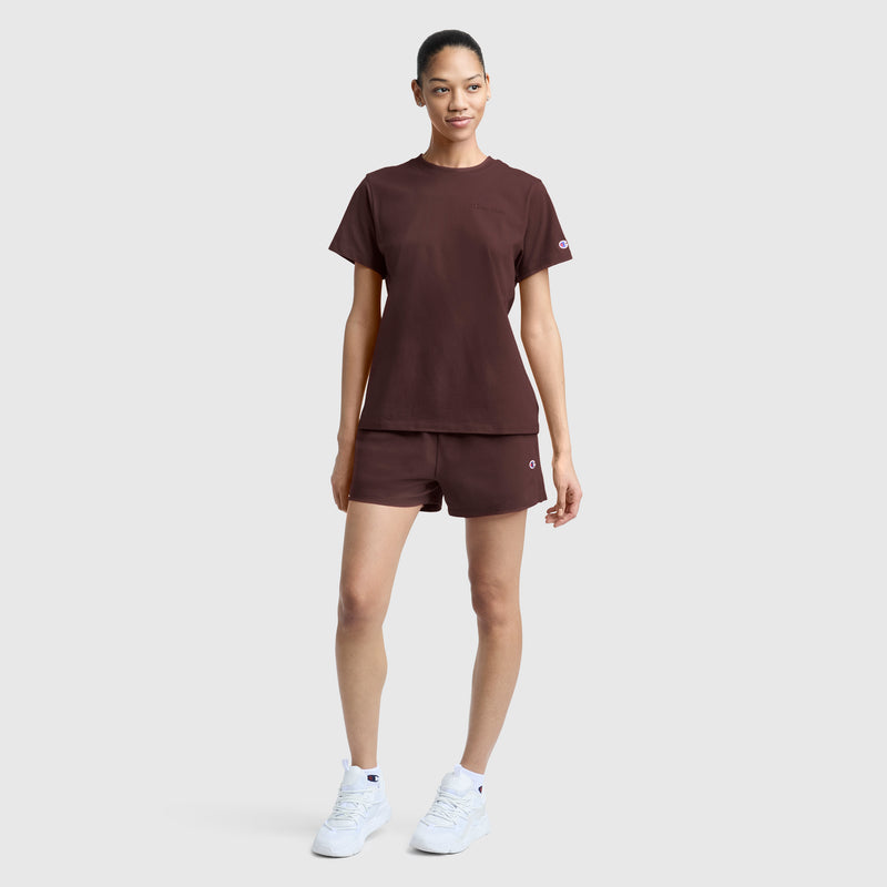 A woman stands against a plain background, wearing the Champion Practice Short in a matching maroon set with a t-shirt and white sneakers. Her hair is tied back, and she looks ahead with a neutral expression.