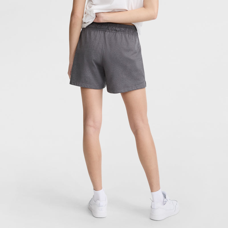 A person stands with their back to the camera, wearing a white t-shirt, Champion 4" Lightweight Lounge Shorts with C Logo in gray, white socks, and white sneakers against a plain white background.