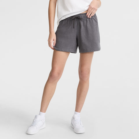 A person is seen from the waist down, wearing a white T-shirt, Champion 4" Lightweight Lounge Shorts with C Logo in gray featuring a drawstring, paired with white crew socks and white sneakers, standing against a plain white background.