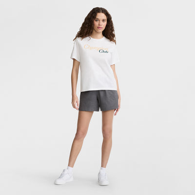 A woman with curly brown hair wears a white T-shirt with Champions Club text, Champion 5" Lightweight Lounge Shorts with a C logo in dark gray, and white sneakers, standing against a plain white background.