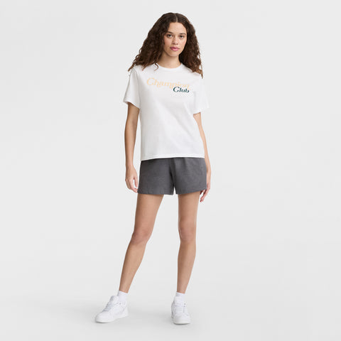 A woman with curly brown hair wears a white T-shirt with Champions Club text, Champion 5" Lightweight Lounge Shorts with a C logo in dark gray, and white sneakers, standing against a plain white background.