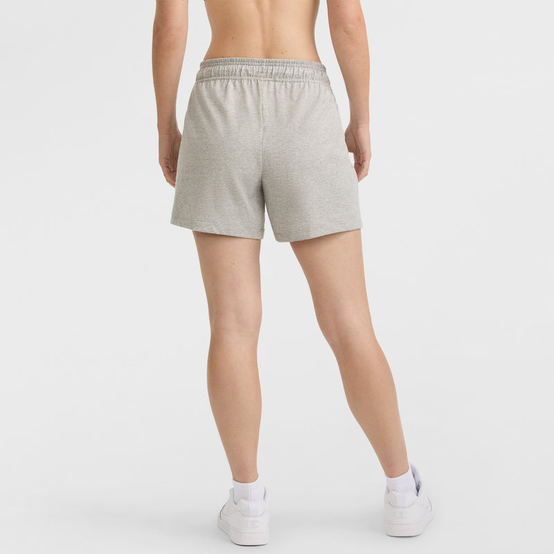 A person stands facing away, wearing Champion 4" Lightweight Lounge Shorts with a C Logo in light gray, paired with white socks and white sneakers. The background is plain white.