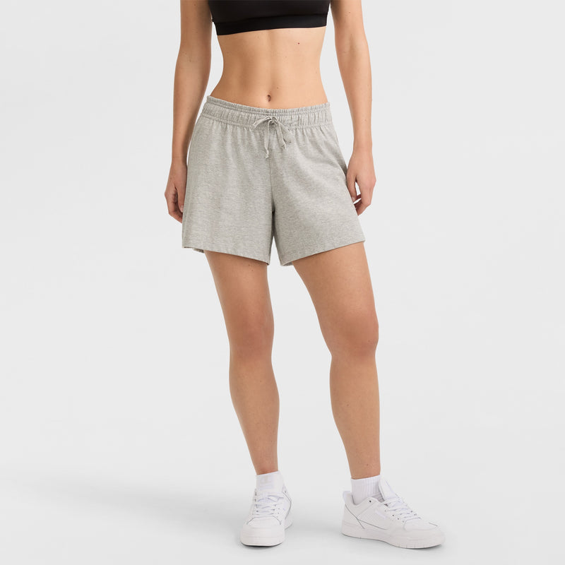 A person wearing a black sports bra, Champion 4" Lightweight Lounge Shorts with C Logo in light gray featuring a drawstring, white socks, and white sneakers stands against a plain white background.