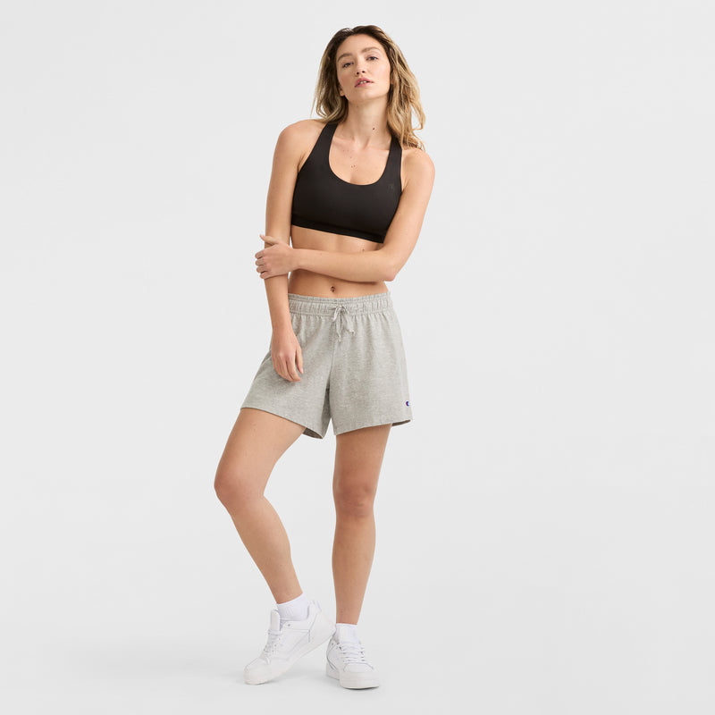 A woman stands posing in a black sports bra, Champion 4" Lightweight Lounge Shorts with C Logo in light gray, and white sneakers against a plain white background.