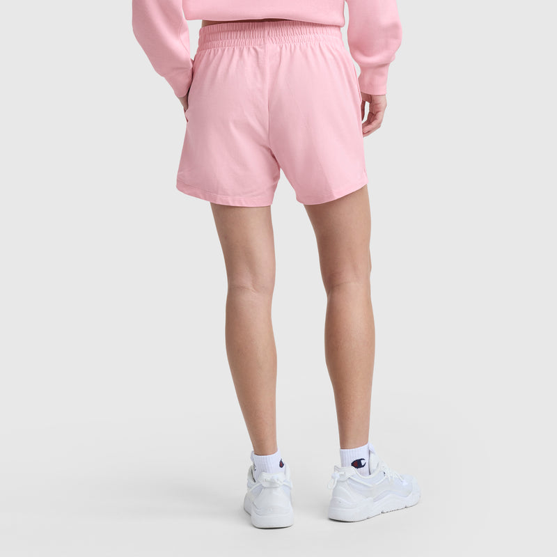 A person stands with their back to the camera, wearing a light pink sweatshirt, Champion 5" Lightweight Lounge Shorts (C Logo), white crew socks with a small logo, and white sneakers against a light, plain background.