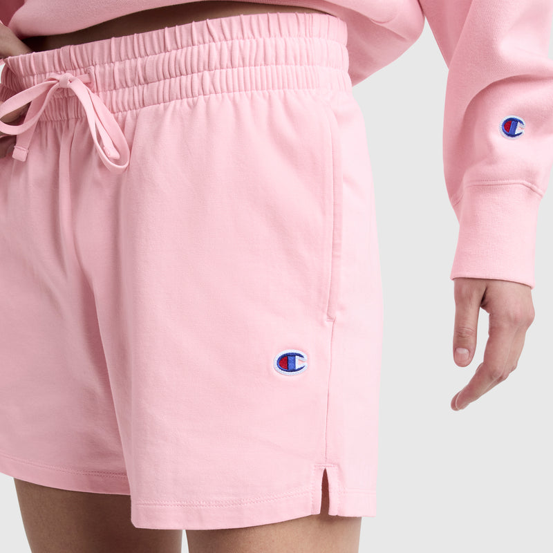 A person wears light pink Champion 5" Lightweight Lounge Shorts, featuring a C logo, elastic waistband with drawstring, and small side slits at the hem, paired with a matching sweatshirt.