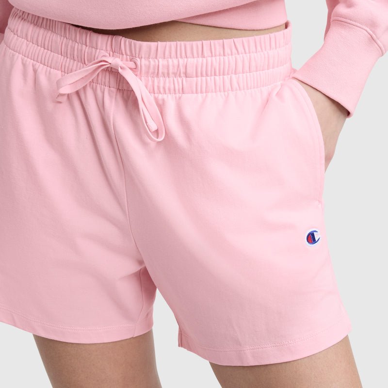 A person wears Champion's 5" Lightweight Lounge Shorts in light pink with a matching sweatshirt, one hand in the pocket. The C logo is visible on the left leg.