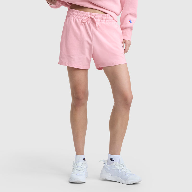 A person in Champion 5" Lightweight Lounge Shorts, light pink, with a matching sweatshirt, white C Logo socks, and white athletic sneakers stands against a plain light background. The person's face is not visible.