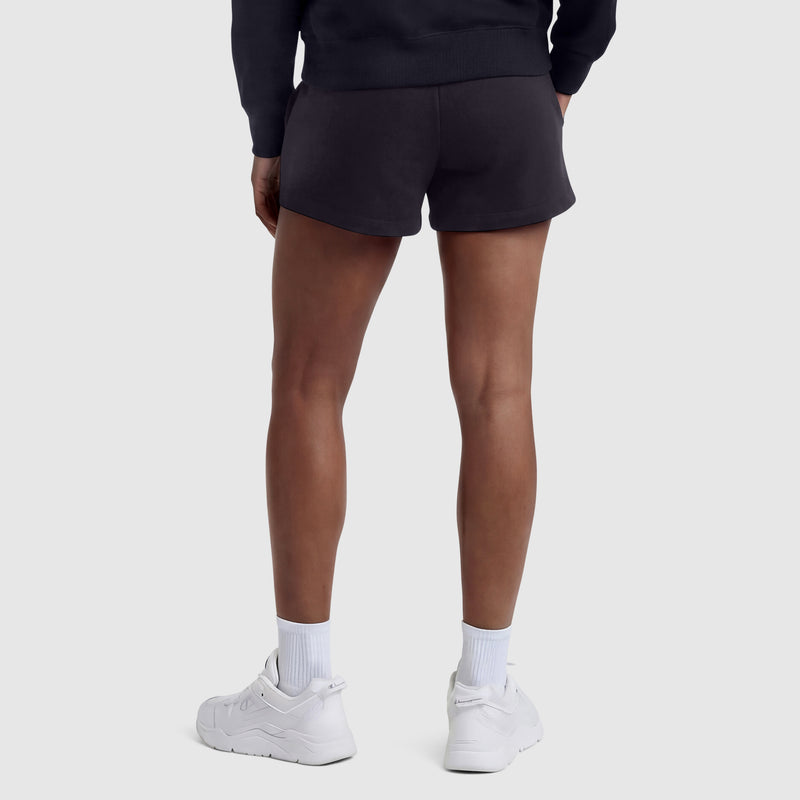 A person stands facing away, wearing a black sweatshirt and Champion 4" Lightweight Lounge Shorts, C Logo, featuring an adjustable waistband, paired with white crew socks and white athletic shoes against a plain light gray background.