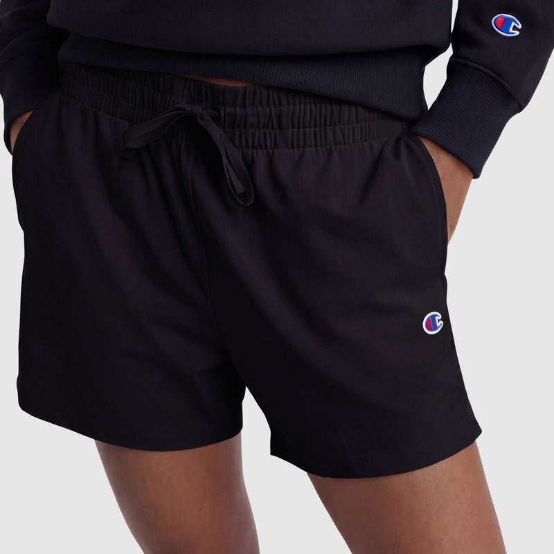 A person wears black Champion 4" Lightweight Lounge Shorts with an adjustable waistband and matching sweatshirt, both featuring the C logo, hands in shorts pockets against a plain, light background.