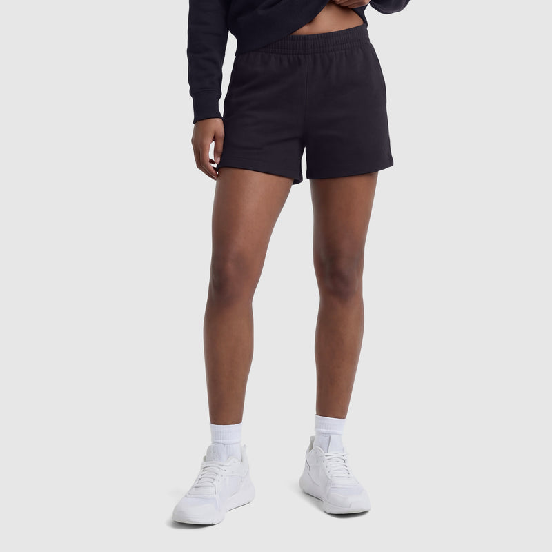 A person stands against a light gray background, wearing black sweatshirt, Champion 4" Lightweight Lounge Shorts with C Logo and adjustable waistband, white crew socks, and white sneakers. The photo is cropped at the shoulders, showing only the lower body.