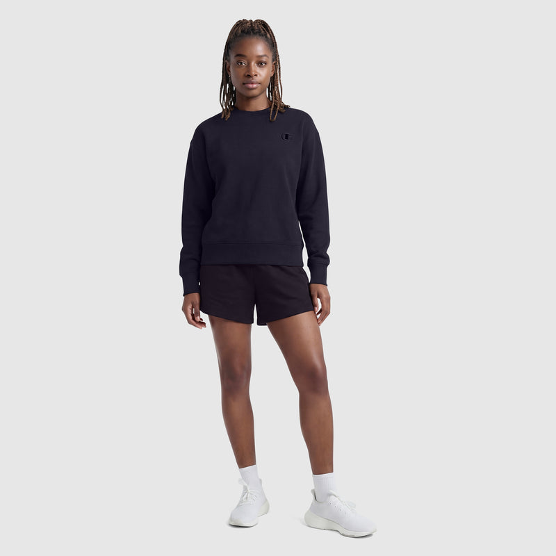 A person stands against a light gray background wearing a black Champion sweatshirt, Champion 4" Lightweight Lounge Shorts with C Logo, white socks, and white sneakers. Their hair is braided and they have a relaxed, neutral expression.